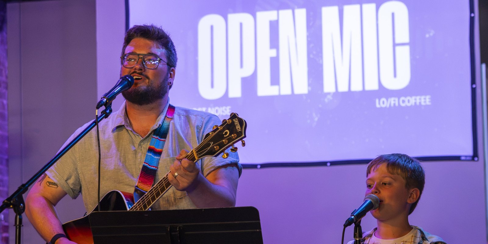A man playing guitar and a boy singing into a microphone under the OPEN MIC projection at Lo-Fi Coffee Bar